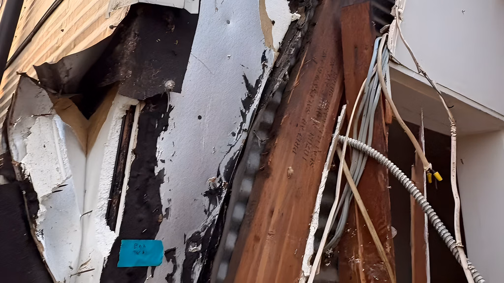 Asbestos-containing black mastic bonded to wood siding and structural framing, with a sample marker taped to the wall